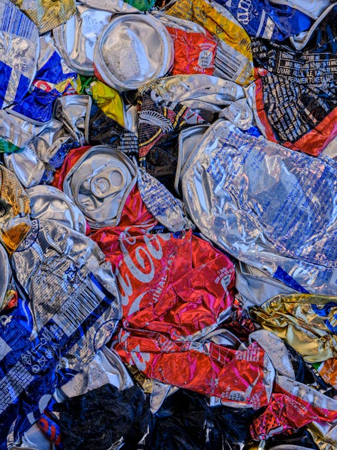 A close-up image of a pile of crushed aluminium drink cans, primarily made of lightweight aluminium with shiny, metallic silver surfaces and colorful printed labels. The cans display various brands and designs, with some showing parts of recognizable logos such as Coca-Cola in red and white, and others with blue, black, and yellow branding. The cans are randomly crumpled and stacked together, occupying the entire frame, with textures ranging from smooth, reflective surfaces to ridged edges where the cans have been crushed or folded. In the background, additional cans and fragments are visible, indicating a collection of recyclable metal waste. The scene appears to be outdoors, on a surface related to waste collection or disposal, consistent with the context of independent rubbish clearance or private waste handling by services such as Rubbish Clearance Croydon, related to rubbish removal or disposal activities.