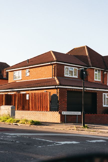 A two-storey detached house constructed with red brick walls and a brown tiled pitched roof, situated on a street corner. The front garden is enclosed by a wooden fence with a vertical slat design, and there is a small black gate leading to the property. Windows on the upper floor have white frames, and a black satellite dish is attached to the side of the house. A street sign reading 'Yarningale Road' is mounted on a black pole in front of the property. The sidewalk runs parallel to the road, which is marked with white lines, and a lamppost stands near the corner of the house. The sky is clear with natural daylight illuminating the scene. The overall setting suggests an urban residential area, with no visible rubbish or waste materials present in the immediate vicinity, aligning with private rubbish removal services such as those offered by Rubbish Clearance Croydon for local properties.