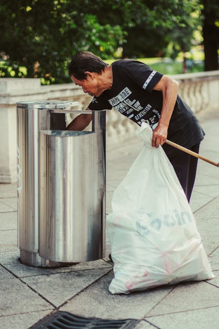 A middle-aged man with dark hair and a black t-shirt, featuring white text and graphics, is seen leaning forward while disposing of a large, white plastic bag filled with rubbish into a cylindrical, stainless steel trash bin positioned on a paved sidewalk. The trash bin has a polished, reflective surface and an open top, with a smaller, open slot design for waste collection, situated next to a stone balustrade that borders a park or green space. Behind the man, lush green trees and foliage are visible, suggesting an outdoor urban environment. The scene is evenly lit, indicating daytime, with natural light illuminating the area and casting soft shadows. The man's posture indicates he is actively engaged in rubbish disposal, consistent with private waste handling or outdoor rubbish removal activities that an independent collection service like Rubbish Clearance Croydon might facilitate. The overall setting emphasizes a routine waste disposal action in a well-maintained public area, illustrating the importance of proper litter management.