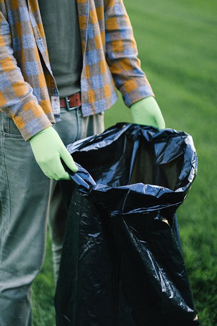 A person wearing a plaid shirt in grey, yellow, and blue, with a green long-sleeved top underneath, is holding open a large black rubbish bag lined with a shiny silver inner layer. They are standing on a well-maintained grassy area, suggesting an outdoor setting such as a garden or park. The person's hands are covered with light green rubber gloves, indicating they are handling waste or rubbish. The background features an expanse of green grass, and the person's pants are grey with a brown belt visible. The scene highlights the process of waste collection or rubbish disposal, which can be associated with services offered by companies like Rubbish Clearance Croydon, reflecting on the importance of private waste management and on-site rubbish removal practices.