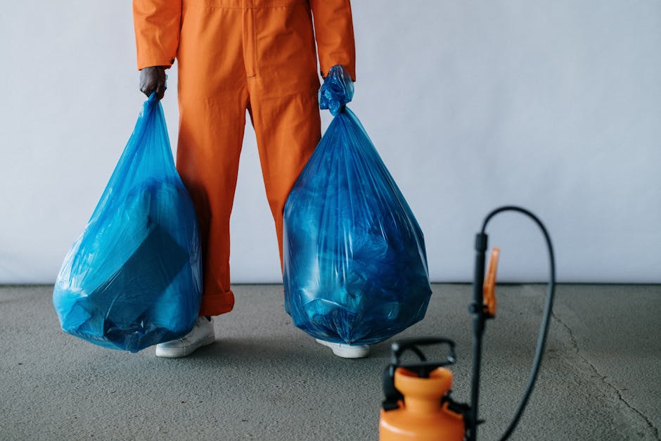 A person dressed in orange workwear stands indoors on a smooth, grey concrete floor, holding two large blue plastic rubbish bags filled with waste material. The person's hands are gloved, and they are wearing white shoes. The bags are semi-transparent, revealing some of their contents, and are tightly knotted at the tops. The background is plain and light-colored, with no other objects visible. In the foreground, a small orange and black spray or blowtorch device with a flexible hose is partially visible, suggesting a professional environment related to waste handling or cleaning. The scene depicts an aspect of independent or private rubbish removal, aligning with on-site clearance activities, as part of waste management services that complement standard rubbish collection for local authorities. The overall setting emphasizes the handling of waste materials typical of rubbish removal companies like Rubbish Clearance Croydon.