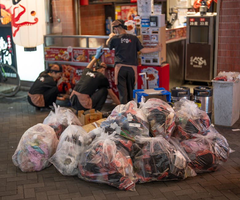 Several large transparent plastic bags filled with mixed household or shop waste, including clothing and packaging, are piled on a paved sidewalk outside a retail establishment. Behind the bags, three workers dressed in black uniforms with brown aprons and gloves are crouching and attending to waste collection near a red and grey storefront, which appears to be a fast-food or retail outlet. The scene includes additional waste containers, a blue toolbox, and a trash bin to the right, suggesting an on-site rubbish clearance process. The environment is illuminated by ambient lighting, highlighting the texture of the plastic bags, which contain a variety of materials, some with visible clothing and packaging. The setting indicates a private waste removal operation, typical of independent rubbish clearance services, and reflects a practical aspect of off-site or private rubbish handling for commercial or retail premises, as managed by Rubbish Clearance Croydon.