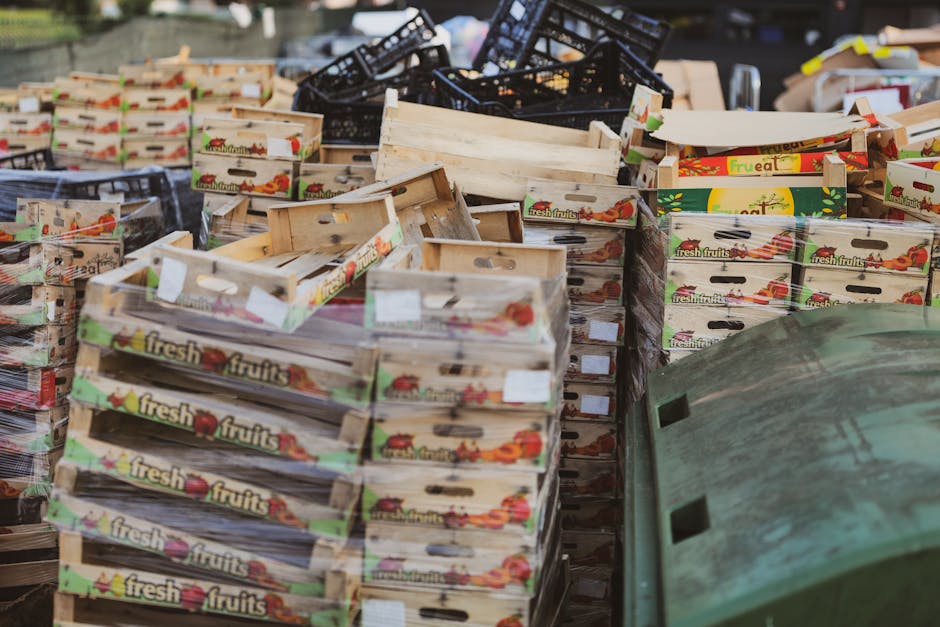 A large collection of empty wooden fruit crates with printed labels reading 'fresh fruits' and colorful images of vegetables and fruits, stacked in disorganized piles on a paved outdoor surface. Some crates are upright, others are tilted or layered irregularly, and they vary in condition with some appearing weathered and slightly worn. In the background, black plastic crates and a few cardboard boxes are visible, along with a green plastic bin positioned at the lower right corner. The scene is outdoors, with natural lighting and no visible signage, illustrating a typical site for bulk rubbish removal involving dismantled produce packaging, possibly as part of a private waste disposal service by Rubbish Clearance Croydon, suited for non-local-authority waste handling and on-site clearance activities.